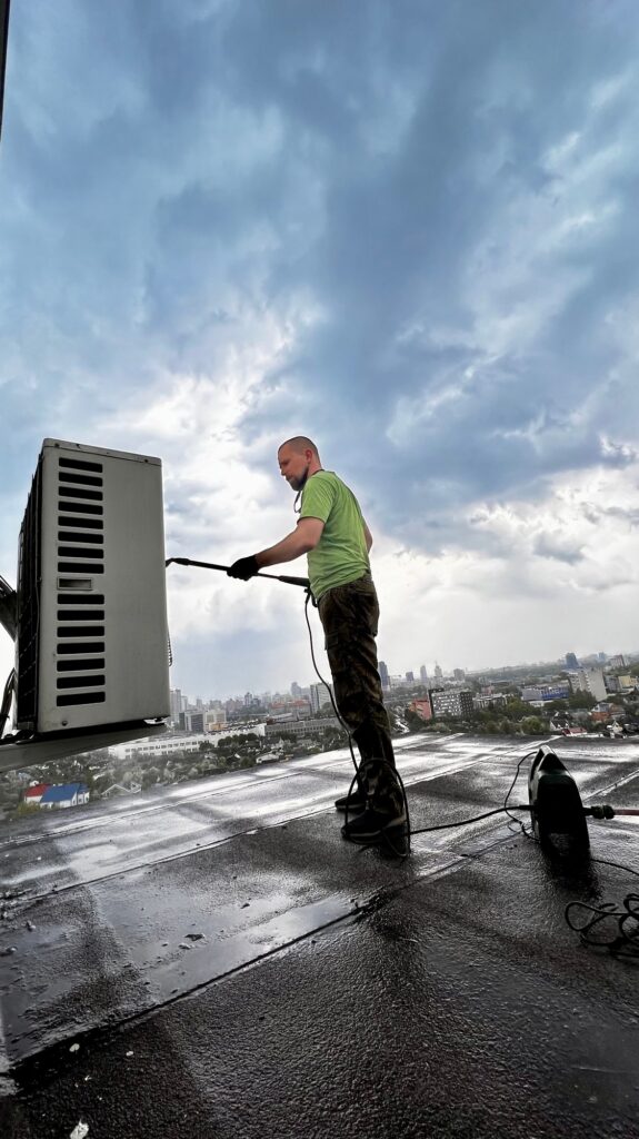 HVAC cleaning services: Technician cleaning an HVAC unit on a rooftop with a pressure washer, city skyline in the background. Phoenix, AZ.