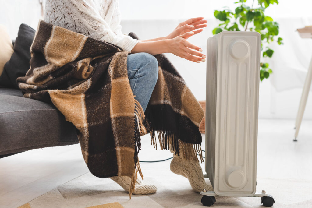 Woman warming hands by space heater with a blanket on couch. Stay warm with heating repair services in Phoenix, AZ.